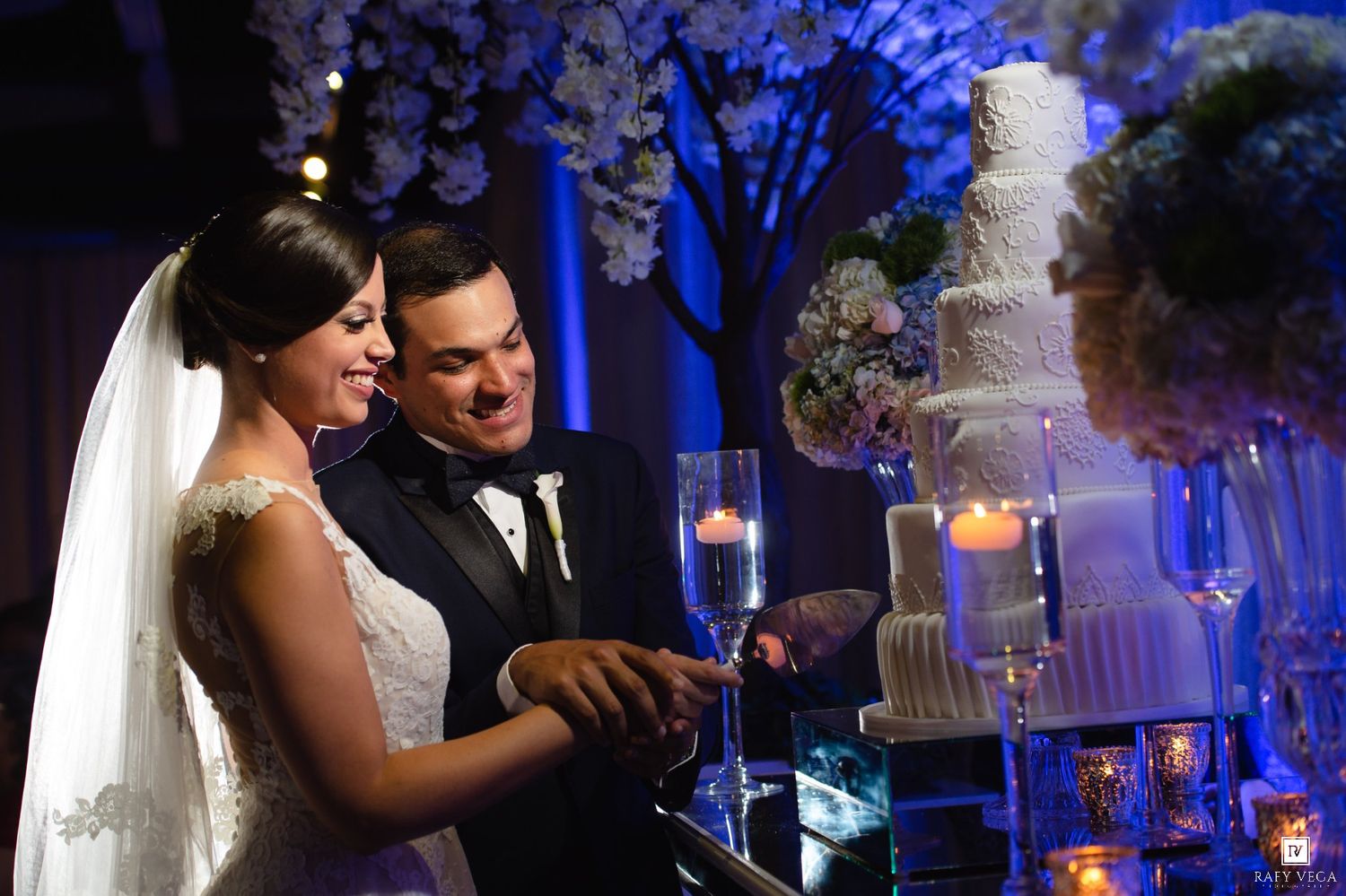 Bride and groom cutting their white wedding cake at elegant reception with blue lighting and floral decor.