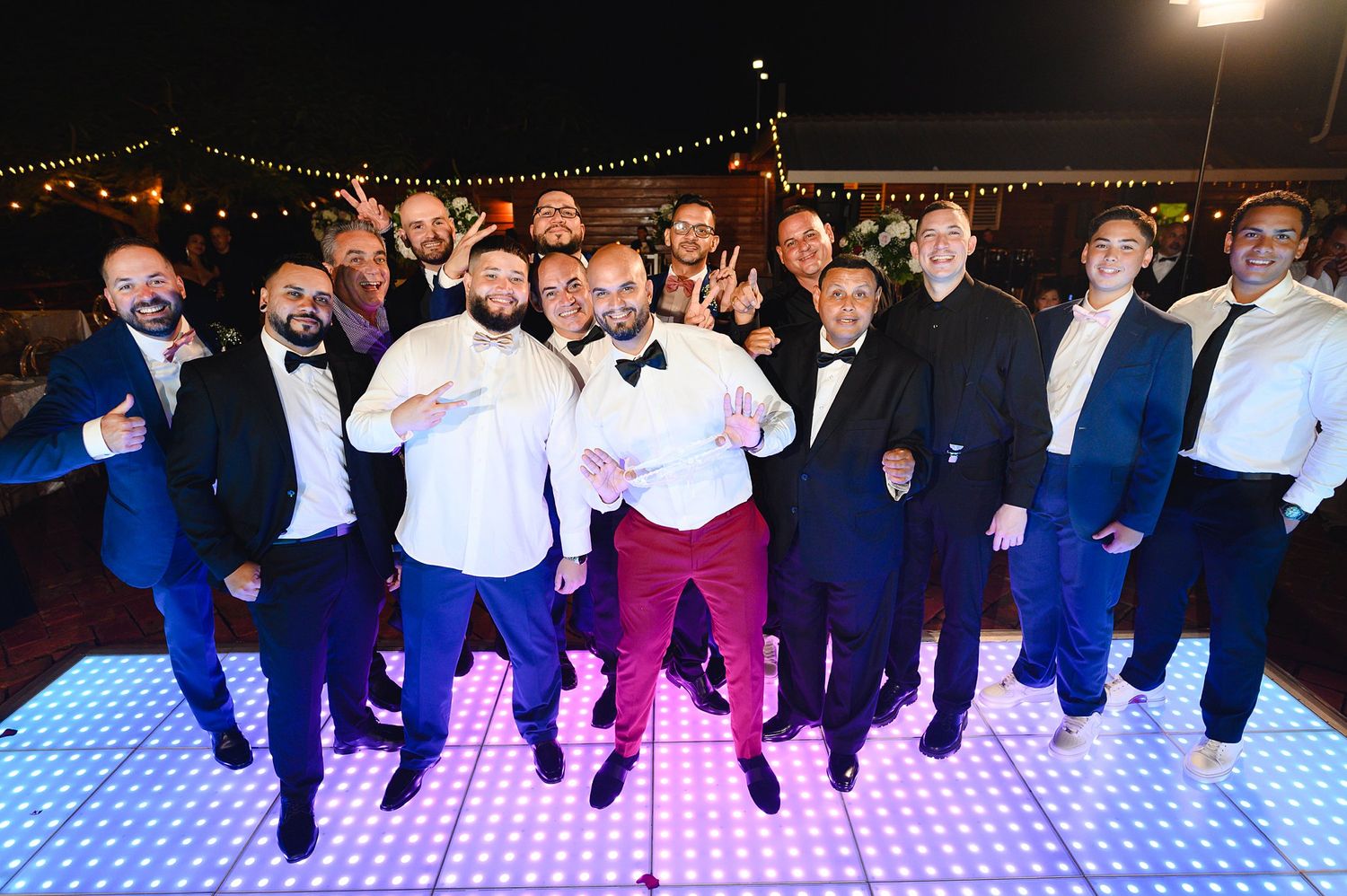Group of men in formal attire posing together on a lit dance floor at an evening celebration.