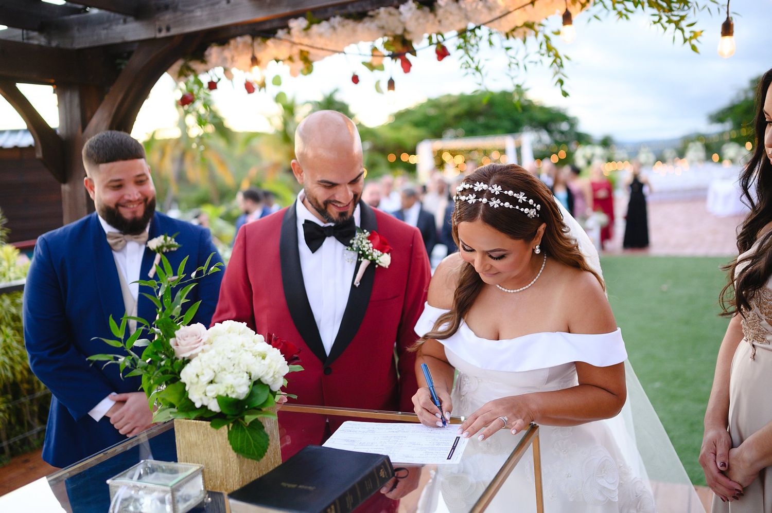 Bride in white gown signs wedding documents beside groom in red tuxedo under pergola at outdoor ceremony.