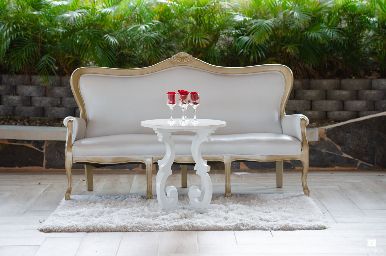 Elegant white vintage sofa and ornate side table with red roses on a rug, set against lush tropical greenery.