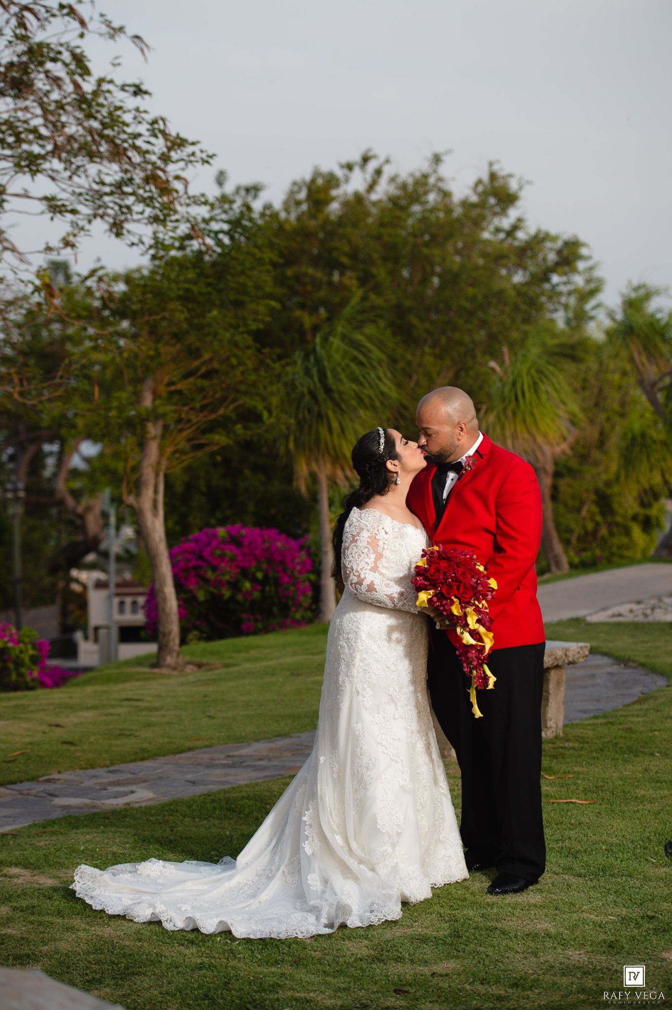 Bride in white lace gown and groom in red jacket share a kiss outdoors on a garden path with tropical trees.