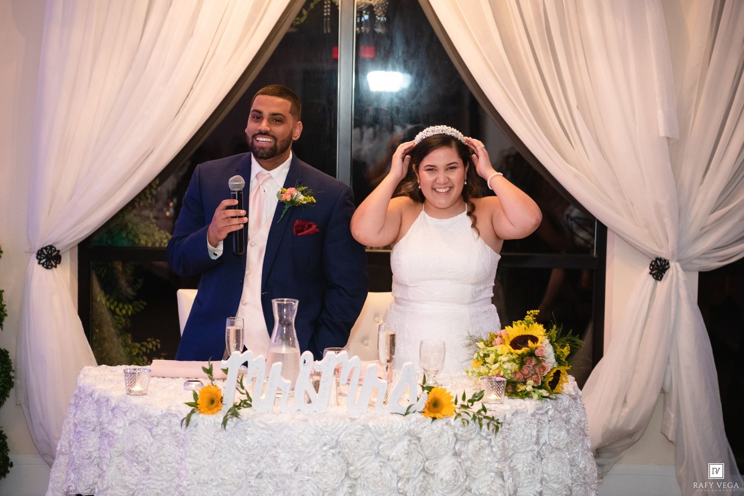 Bride and groom smile at wedding reception head table adorned with sunflowers and white roses.