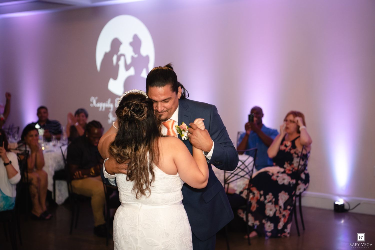 Bride and groom share their first dance at a wedding reception with purple uplighting and guests watching in background.