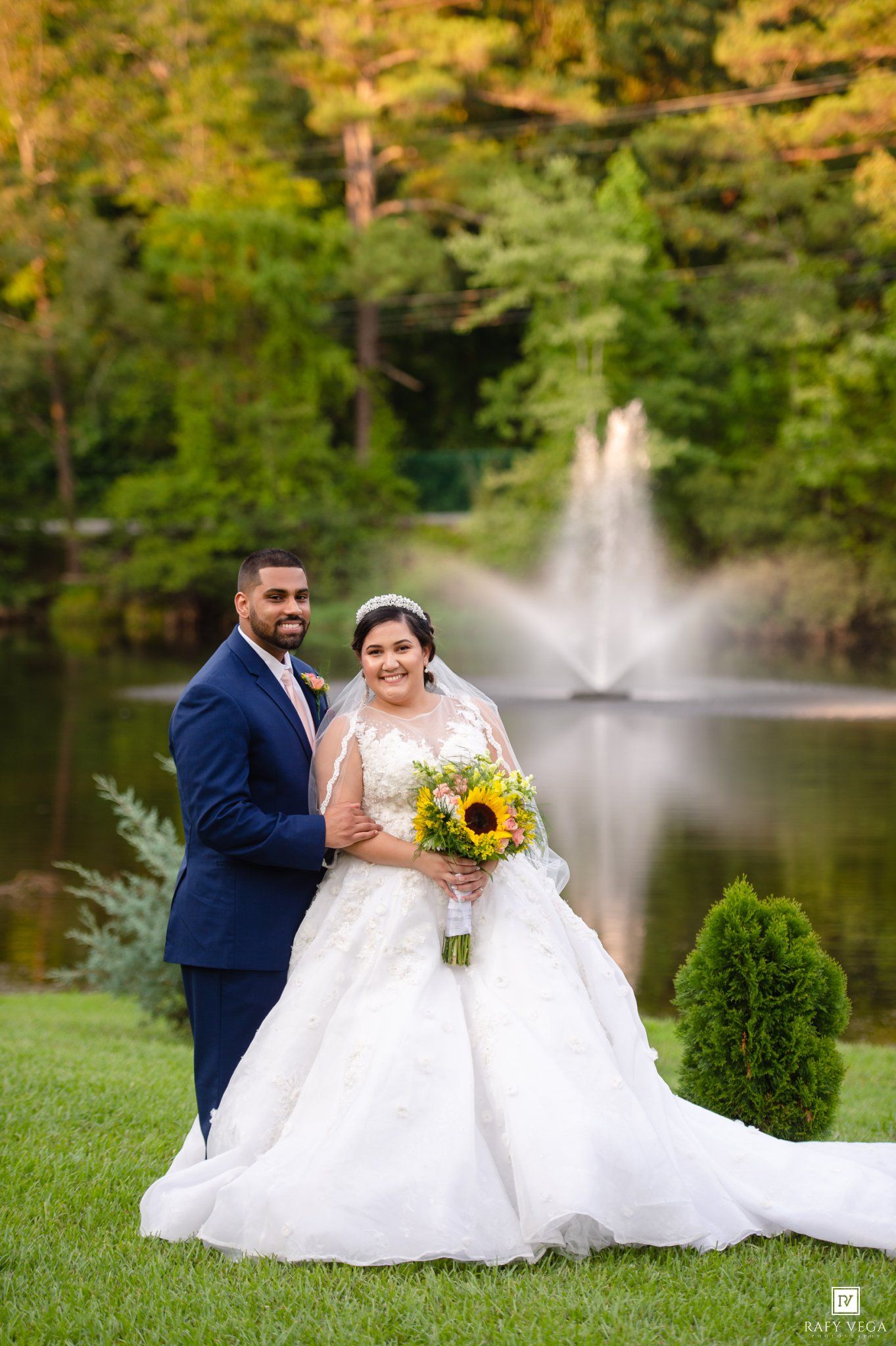 Bride in white ball gown holding sunflower bouquet poses with groom in navy suit by a pond fountain.