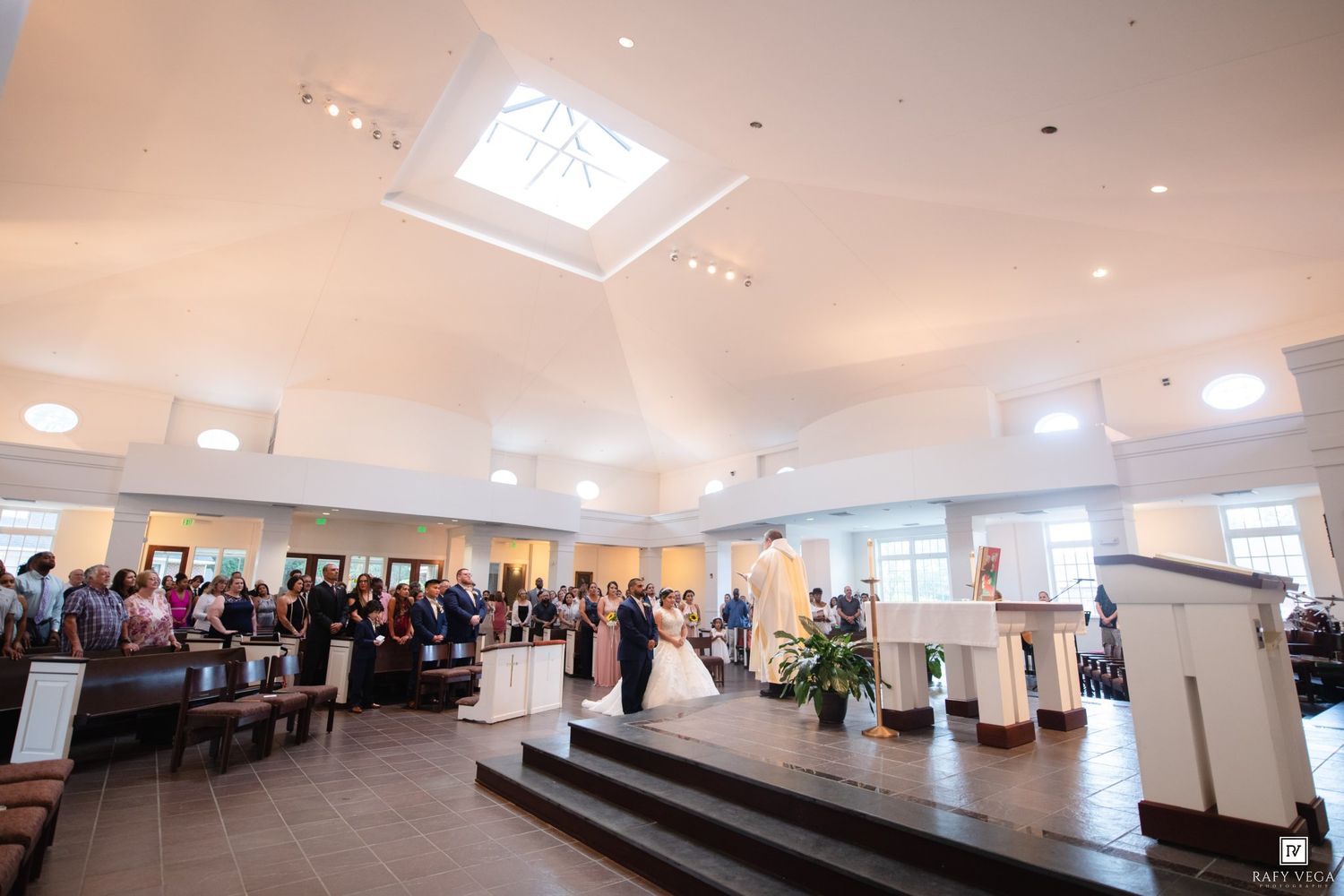Wedding ceremony in a bright church with guests seated in pews, bridal party at the altar under a skylight.