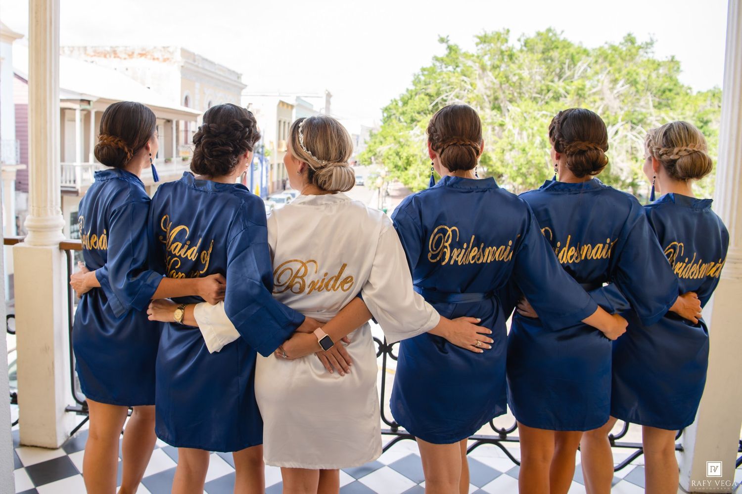 Bride and bridesmaids wearing matching navy and white satin robes with gold script, posing together with backs to camera.
