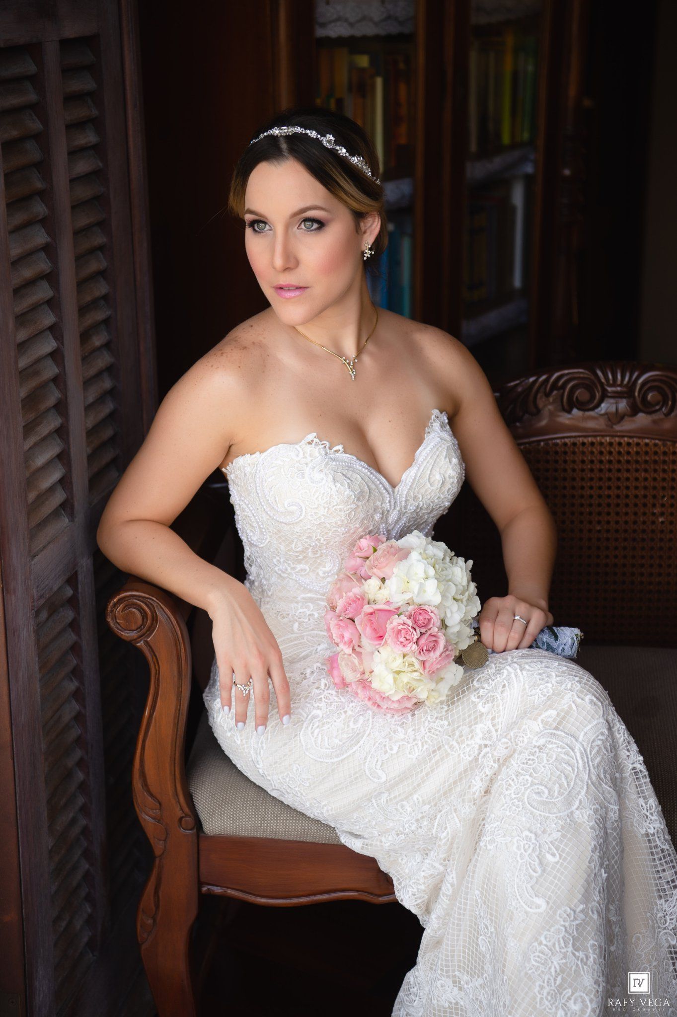 Elegant bride in white lace strapless gown seated in chair holding pink and white rose bouquet with crystal hair accessory.