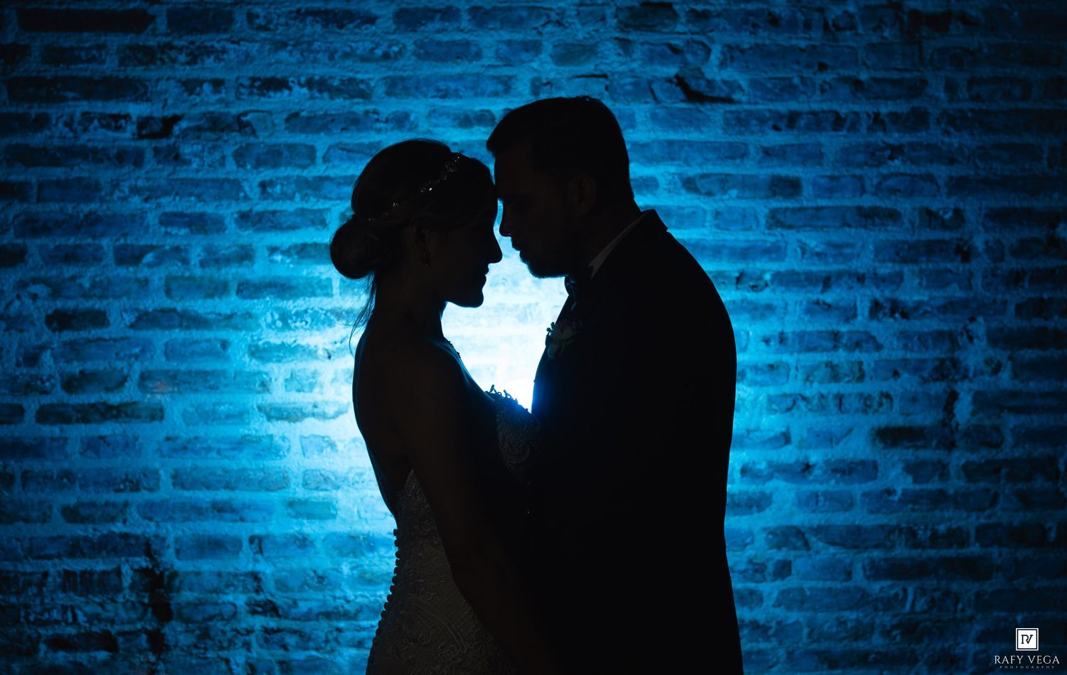 Romantic silhouette of a bride and groom facing each other against a blue-lit brick wall.