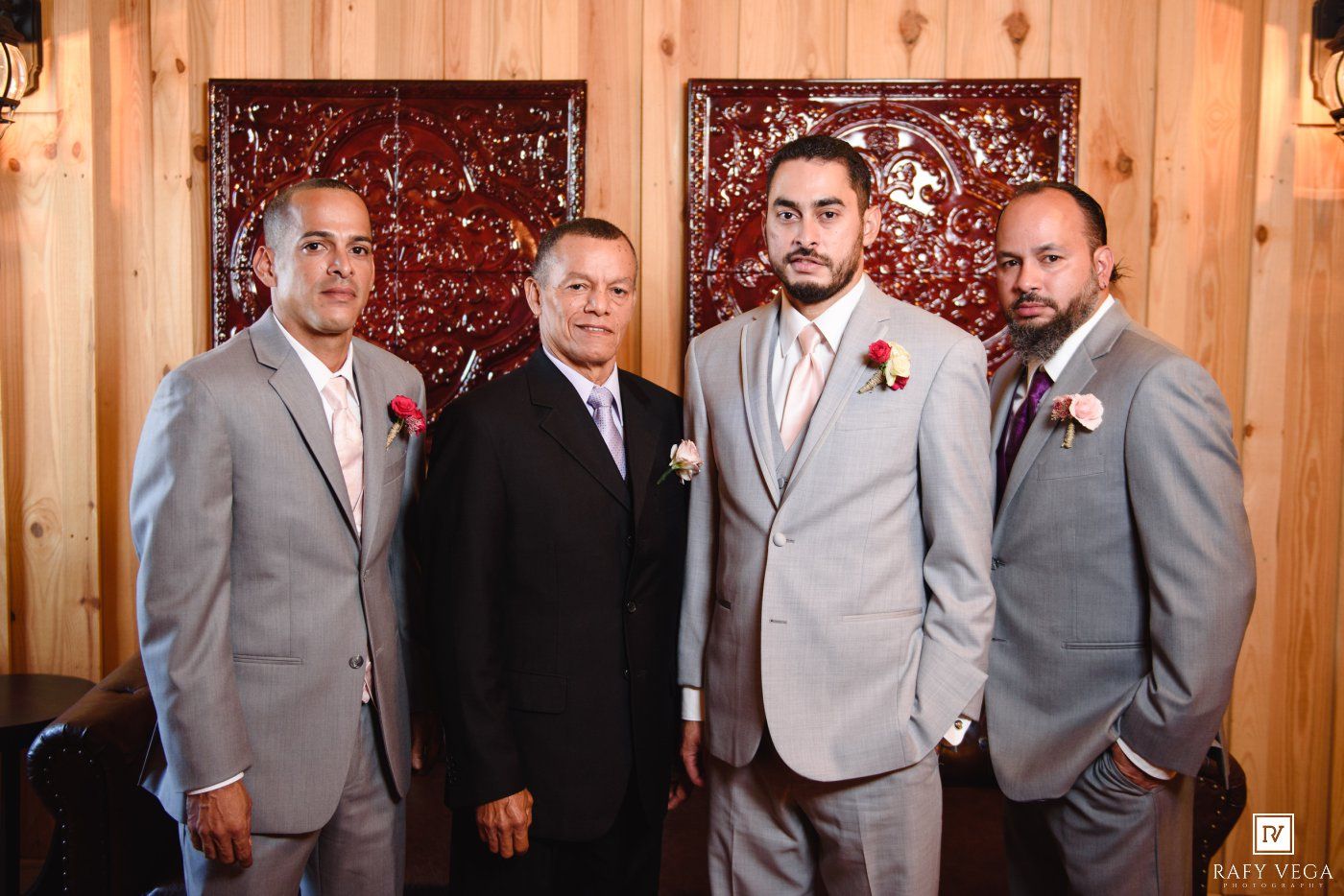 Four men in gray suits with boutonnieres pose together at a wedding venue with wood-paneled walls.