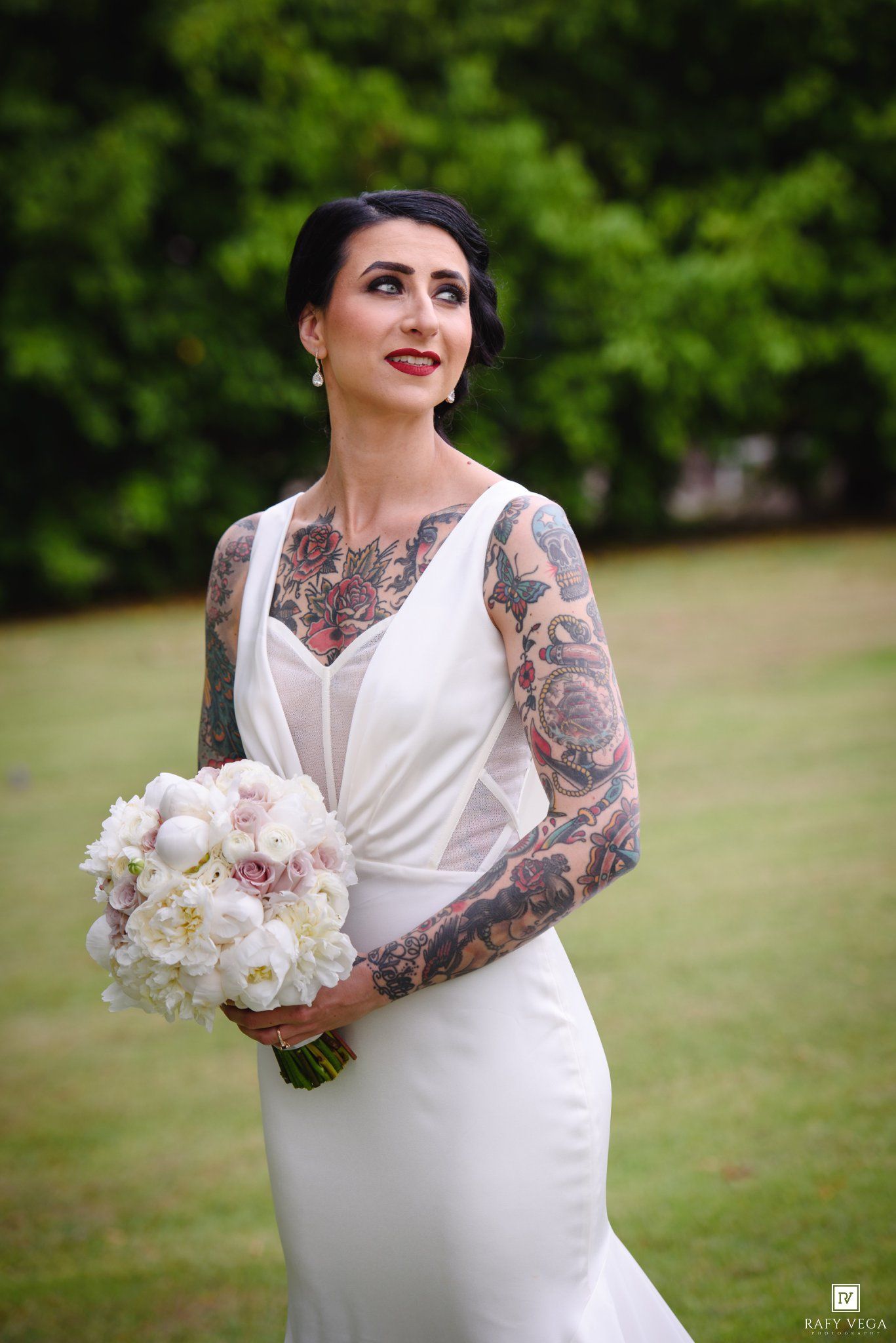 Elegant tattooed bride in sleeveless white gown holding pink and white bouquet looking up outdoors.
