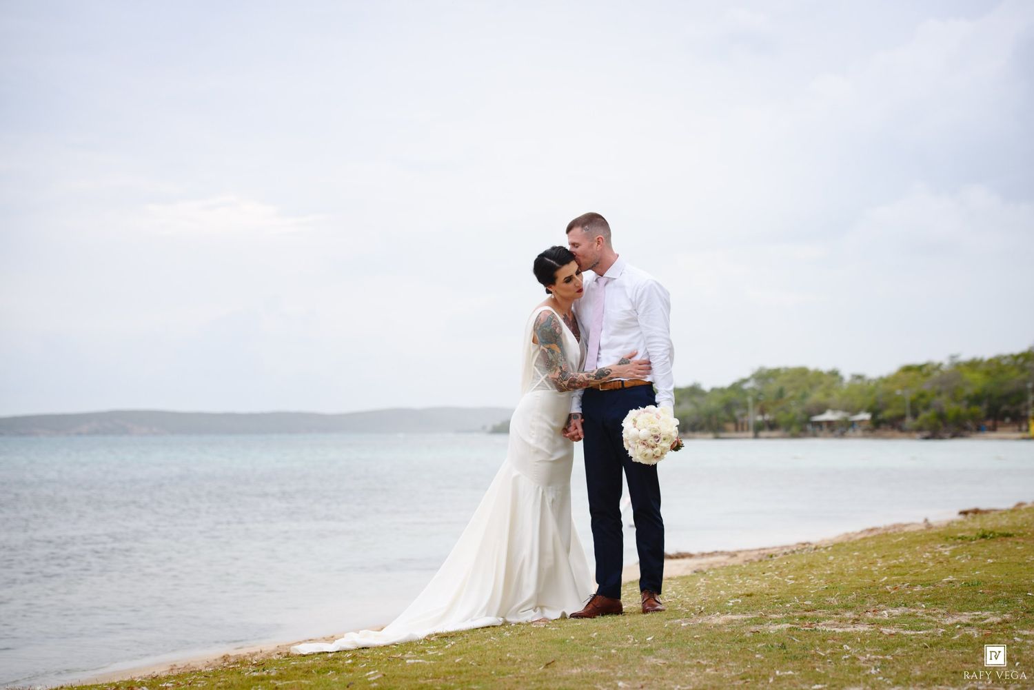 Bride and groom share a romantic kiss by the ocean shore, bride in elegant white gown holding bouquet.