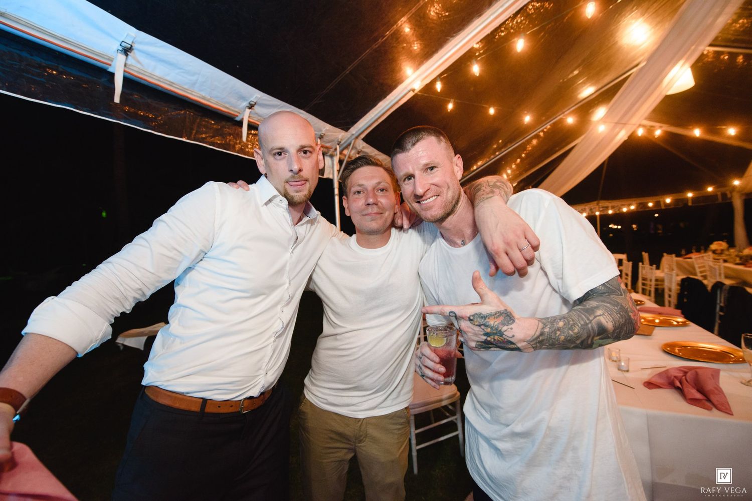 Three men smiling and posing together under string lights at an elegant outdoor wedding reception.