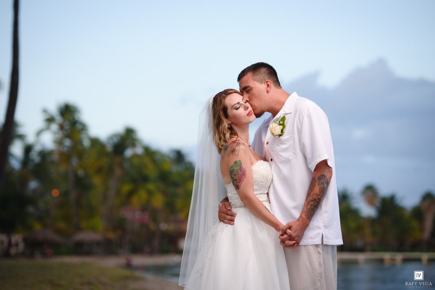 Couple in wedding attire sharing a romantic kiss by a tropical waterfront with palm trees and mountains in the background.