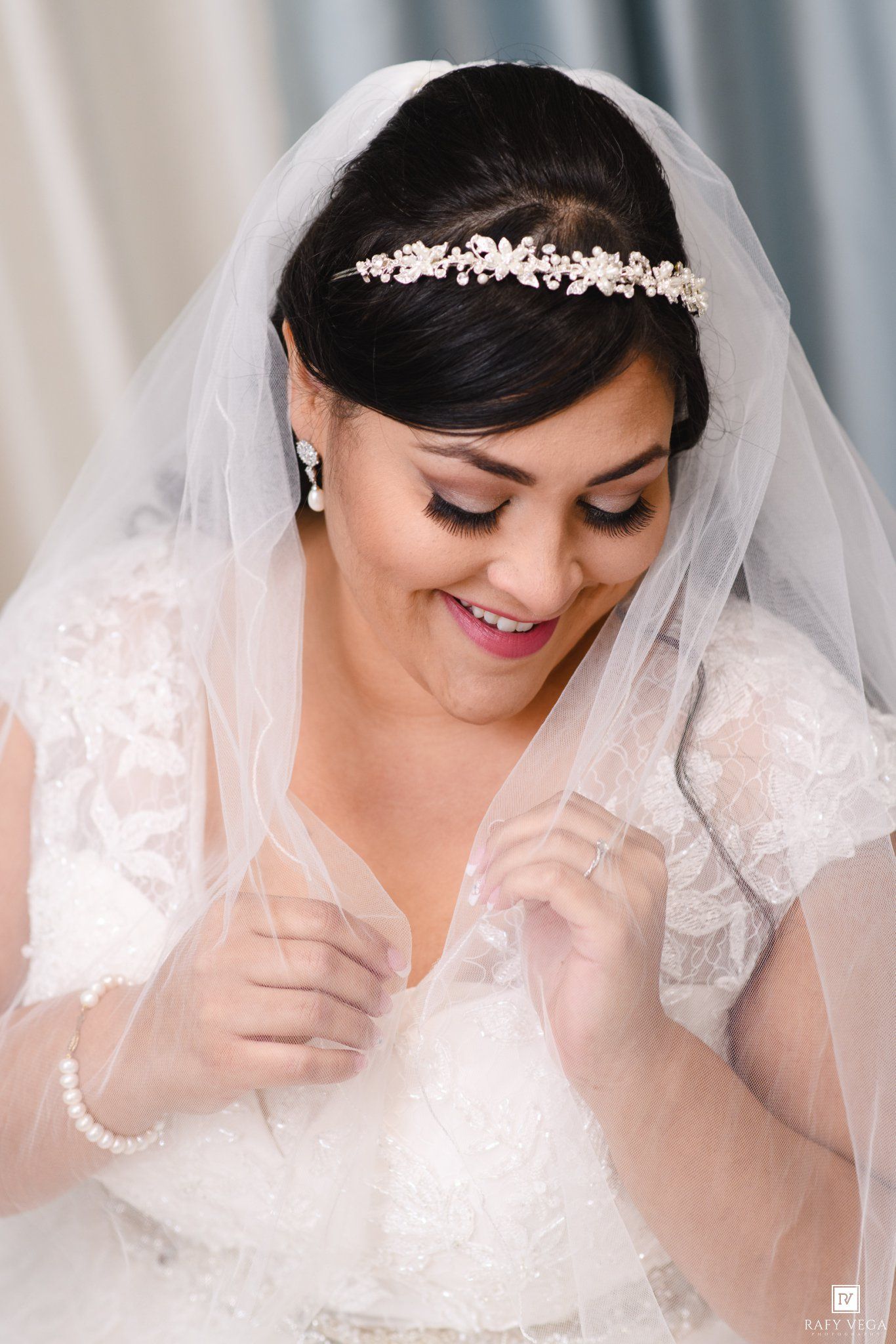 Bride in lace wedding dress and floral headpiece smiles while adjusting her sheer veil.