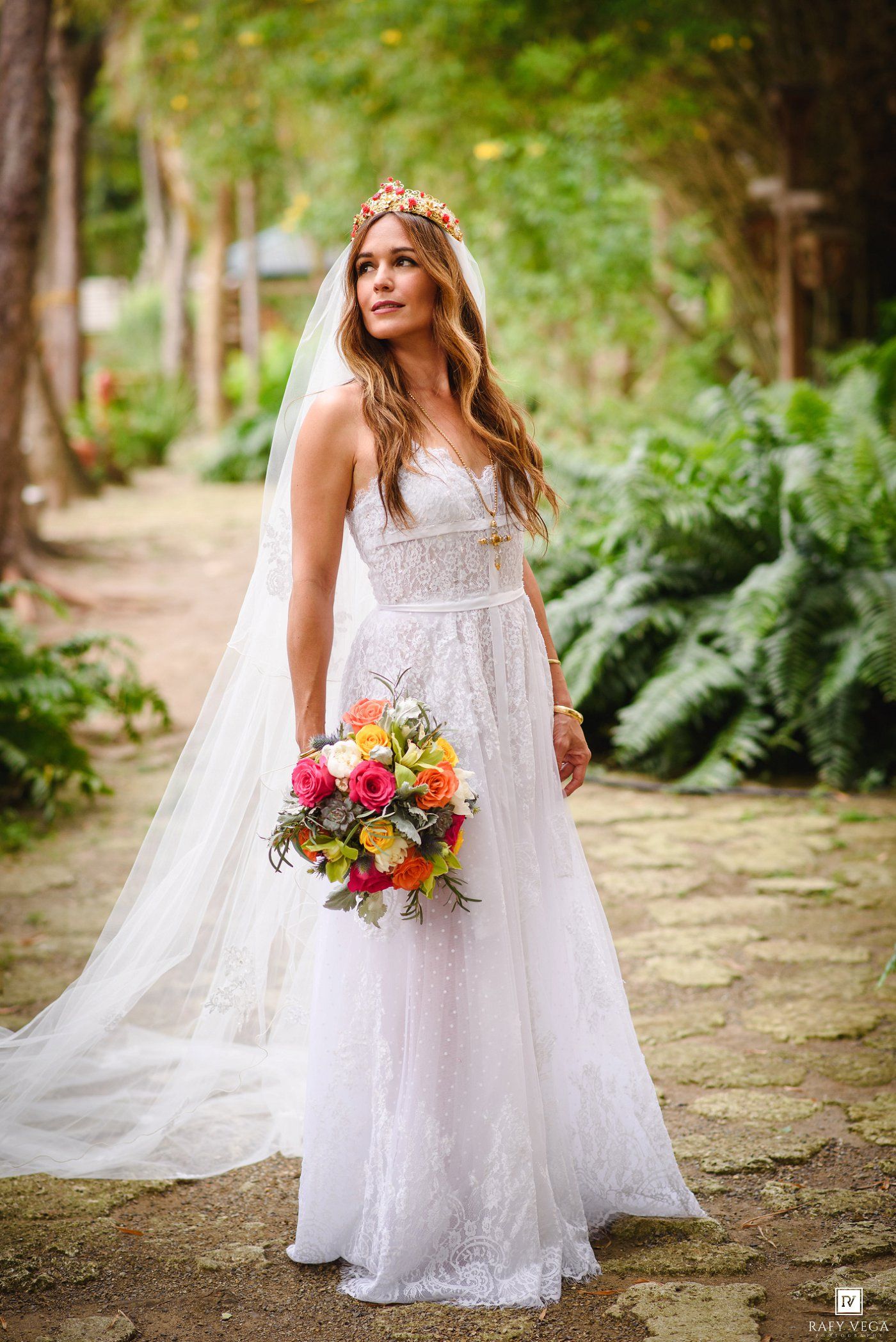 Bride in white lace gown with veil and floral crown holds colorful bouquet on a tree-lined garden path.