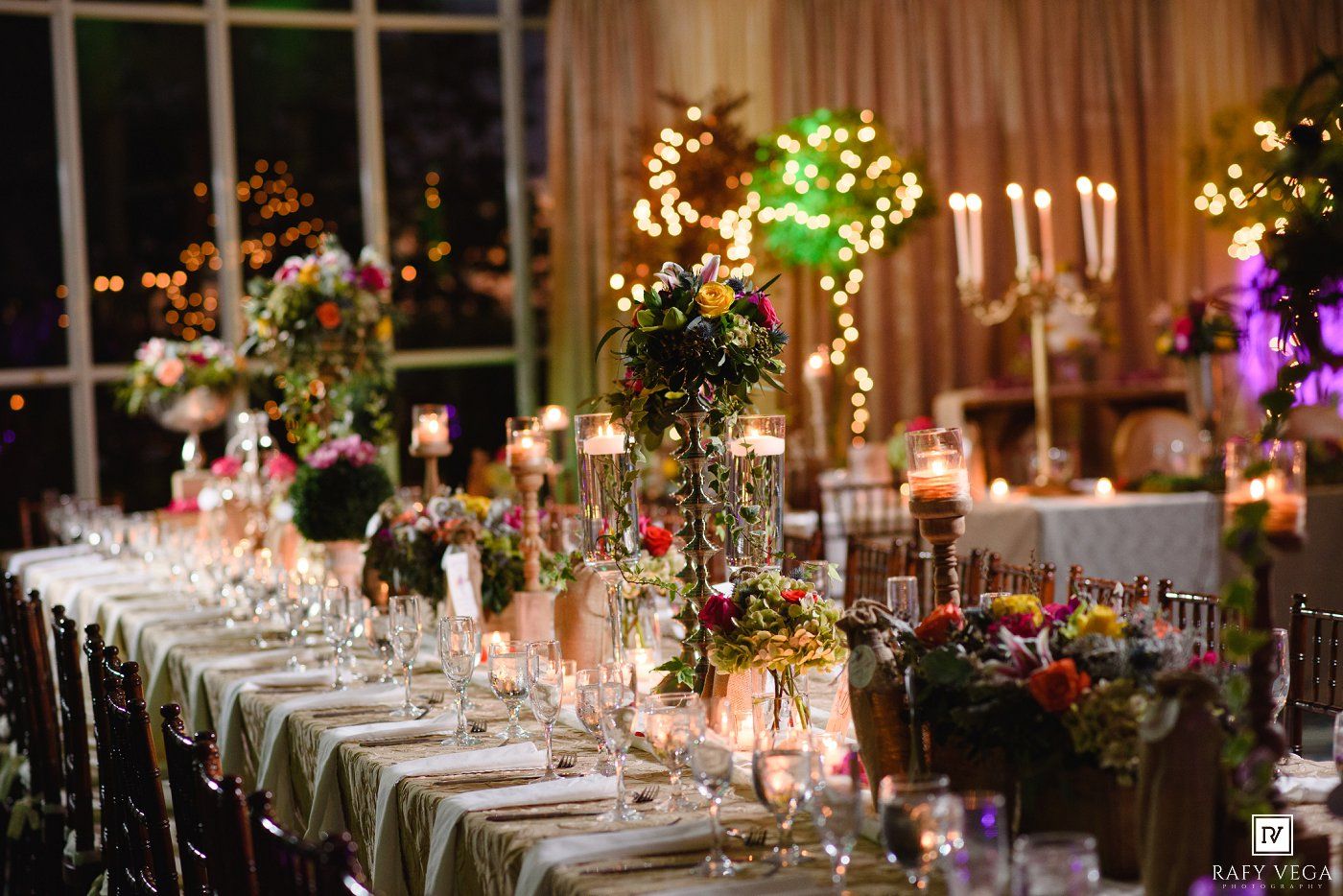 Elegant wedding reception table with tall floral centerpieces, candles, and colorful blooms set against a warmly lit venue.