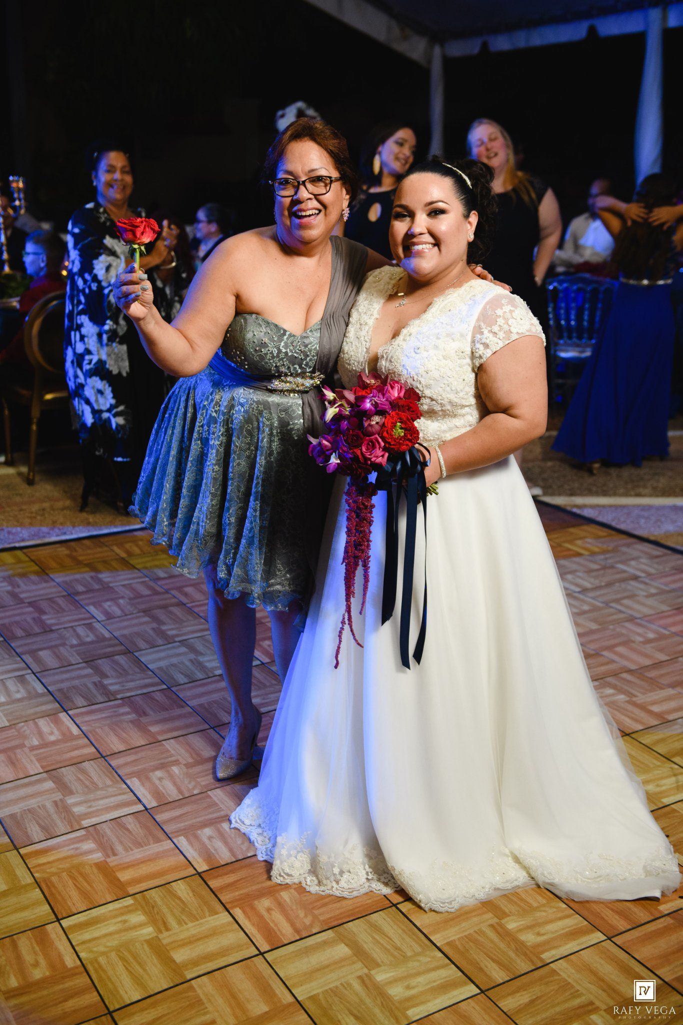 Bride in white lace gown holds bouquet beside smiling woman in blue dress at wedding reception.