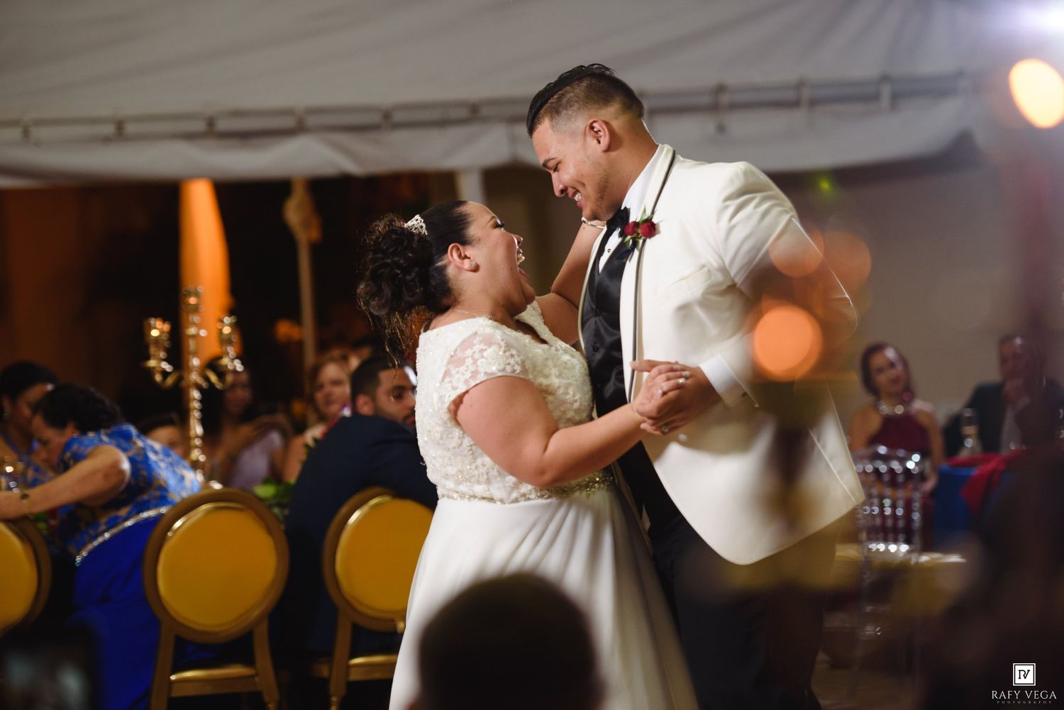 Bride and groom share their first dance at wedding reception, smiling joyfully under warm golden lights.