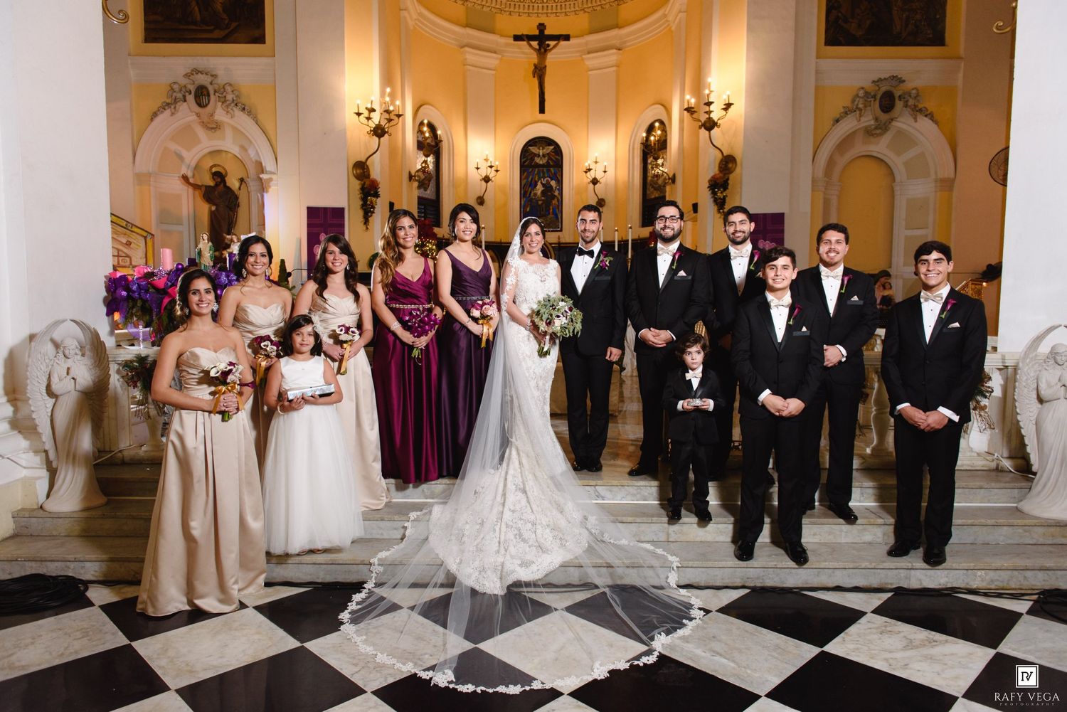 Wedding party poses in elegant venue with checkered floors, bride in lace gown, groom in tuxedo, bridesmaids in burgundy dresses.