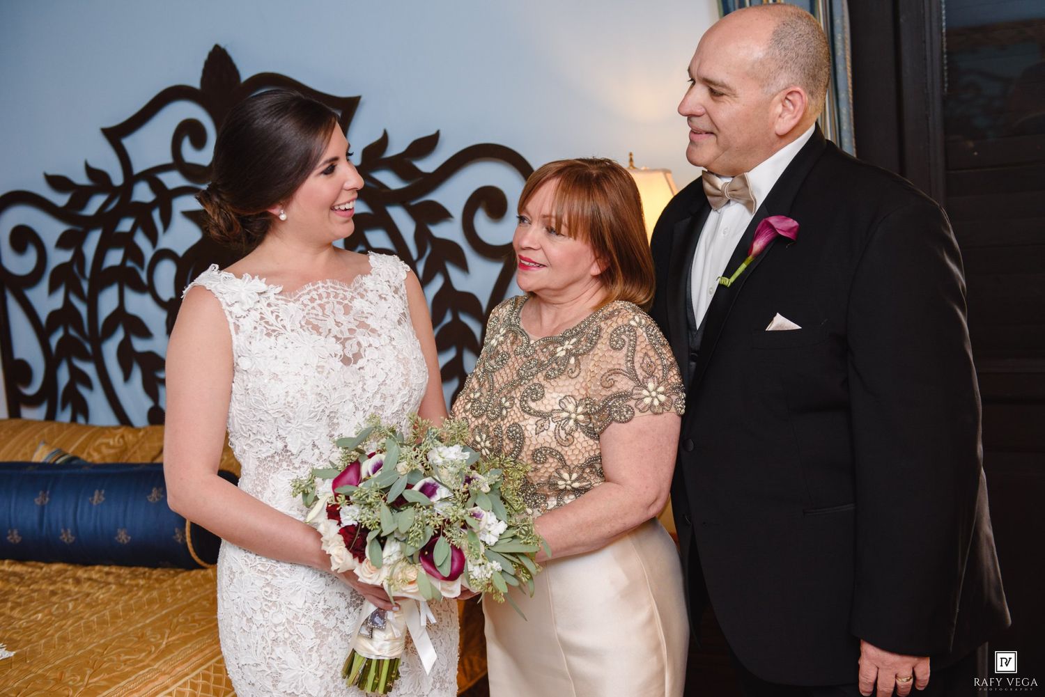 Bride in white lace gown holds bouquet while posing with two guests at an elegant wedding reception.