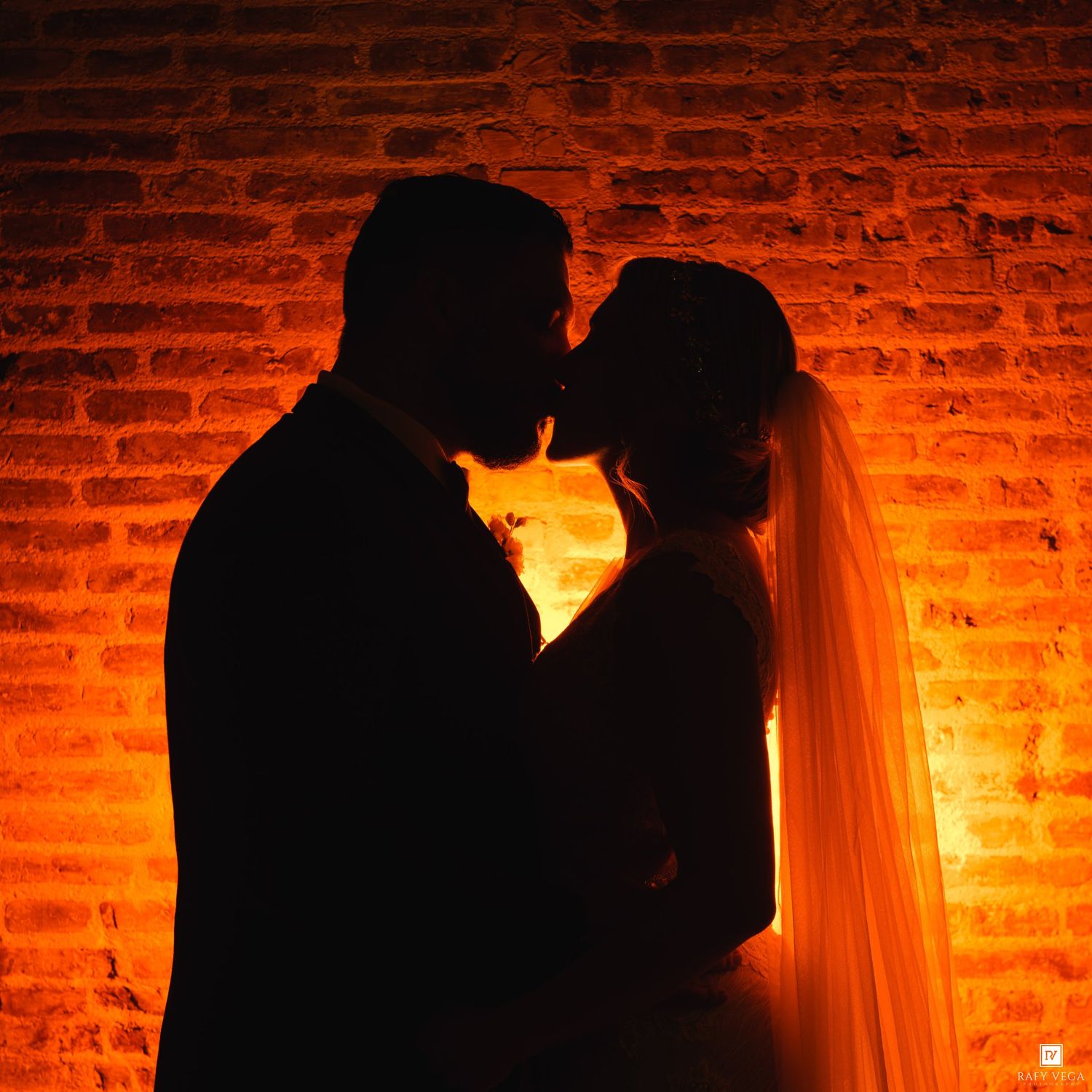 Silhouette of a bride and groom kissing against a warm glowing brick wall with romantic amber lighting.