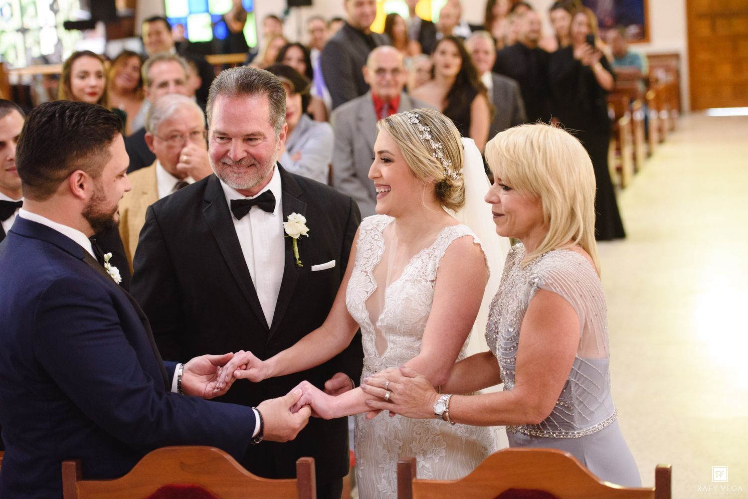 Bride and groom exchange rings at church wedding altar, flanked by smiling parents in elegant formal attire.