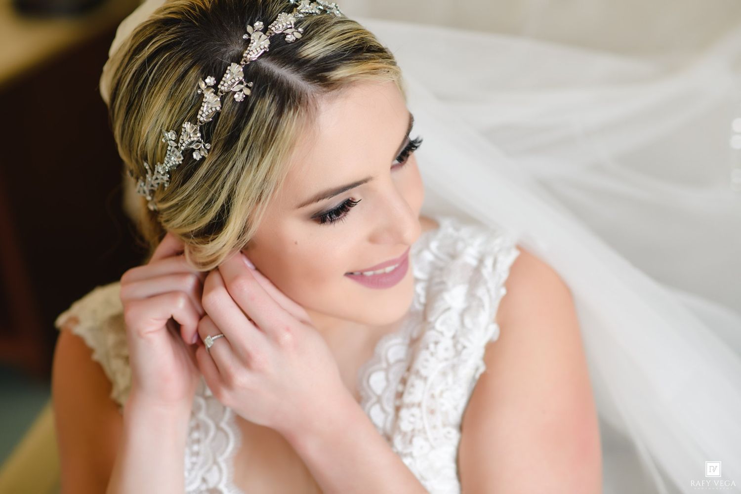 Bride in white lace gown adjusting earring, wearing floral crystal headpiece and veil on wedding day.