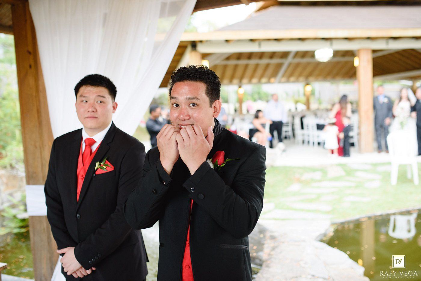 Emotional groom covering his mouth in awe at outdoor wedding ceremony, with groomsman in black suit and red tie behind him.