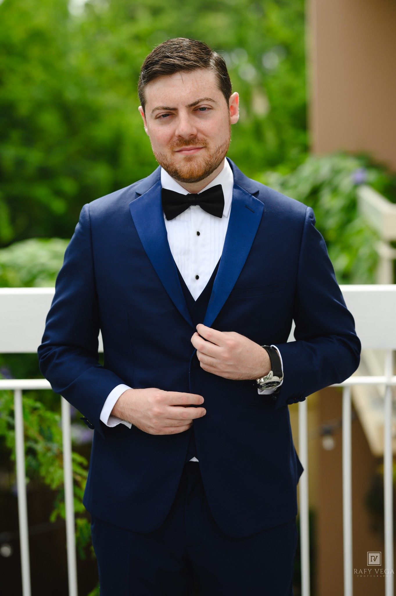 A man in a navy blue tuxedo with black bow tie and vest adjusts his jacket outdoors at a formal event.
