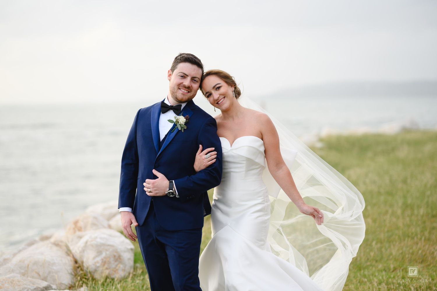 A smiling bride and groom pose together near a rocky shoreline, her veil flowing in the wind on their wedding day.