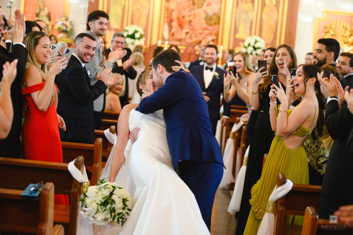 Groom dips bride for a kiss at church wedding altar while guests cheer and photograph the romantic moment.