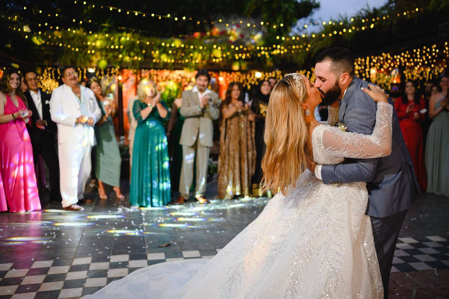 Bride and groom share first dance at elegant outdoor wedding reception with string lights and guests applauding.