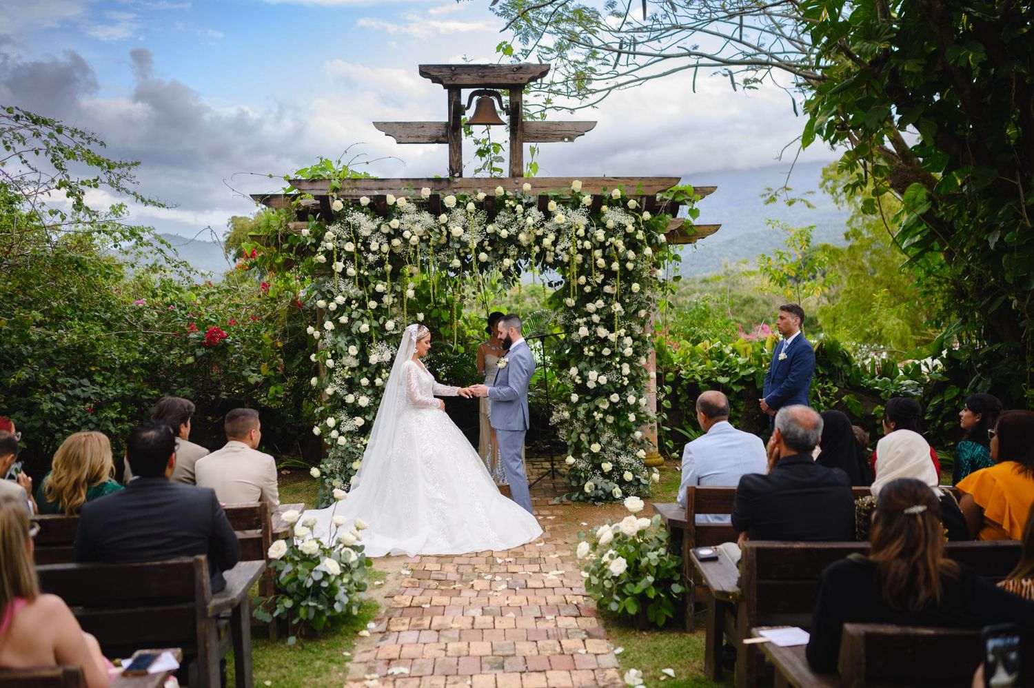 Outdoor garden wedding ceremony with guests seated as couple holds hands before floral arch.