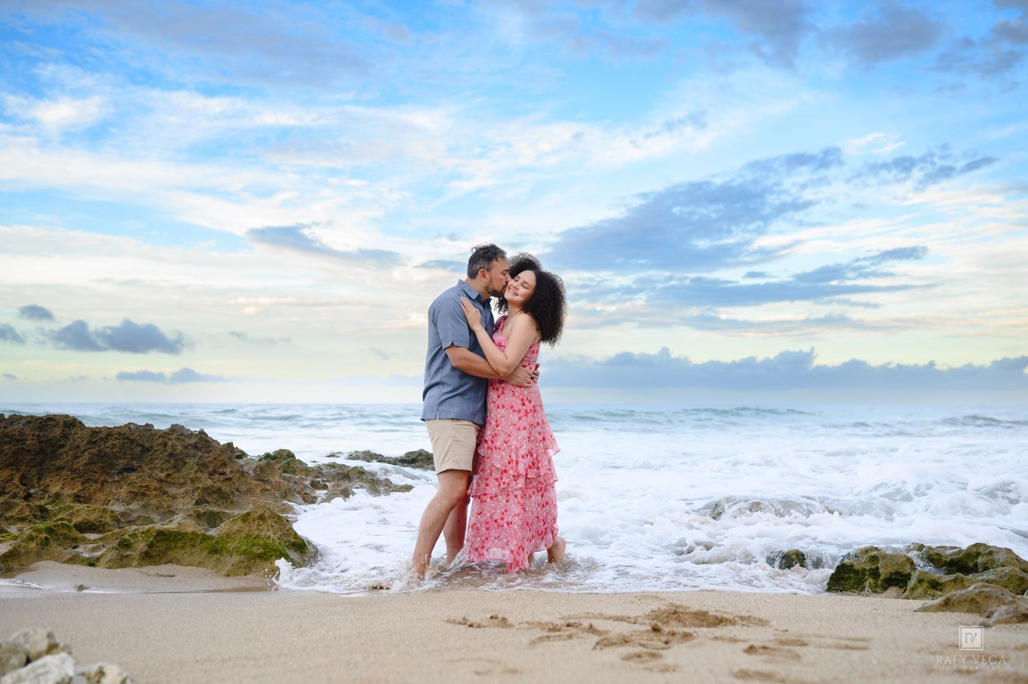 Couple kissing on a tropical beach with ocean waves, rocks, and a beautiful cloudy sky backdrop.