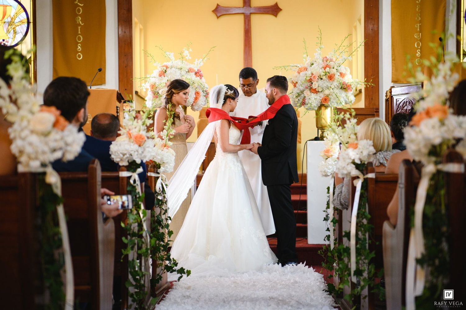 A bride and groom exchange vows at a church altar adorned with white floral arrangements and a large wooden cross.
