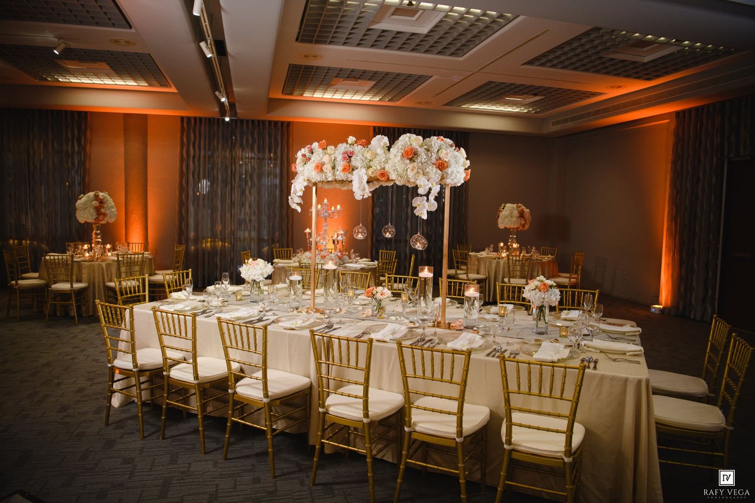 Elegant wedding reception hall with gold chiavari chairs, long tables, tall floral centerpieces, and warm orange ambient lighting.
