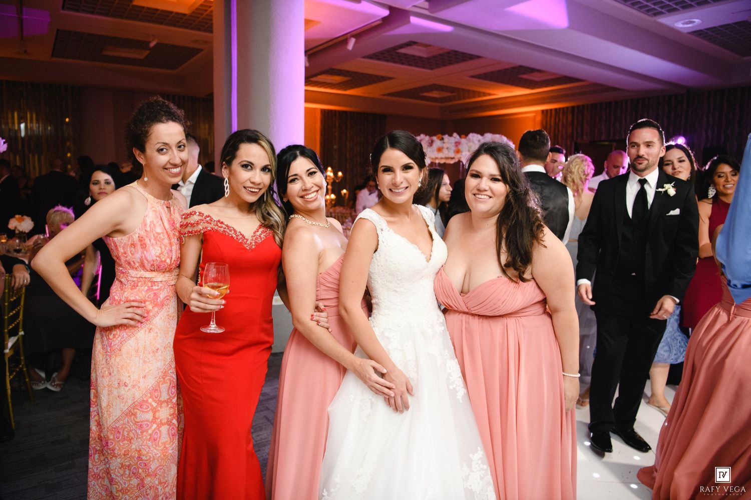 Bride poses with bridesmaids in coral pink dresses at elegant wedding reception venue.