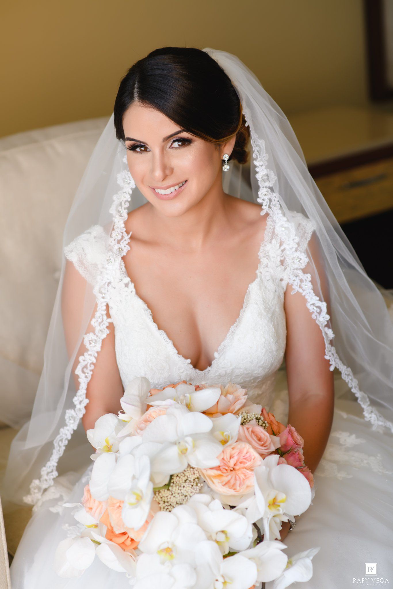 Bride in white lace gown and veil holding white orchid and peach rose bouquet, smiling elegantly on wedding day.