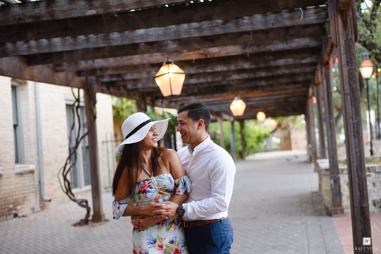 Couple smiling at each other under a rustic pergola with lanterns, woman in floral dress and white hat.