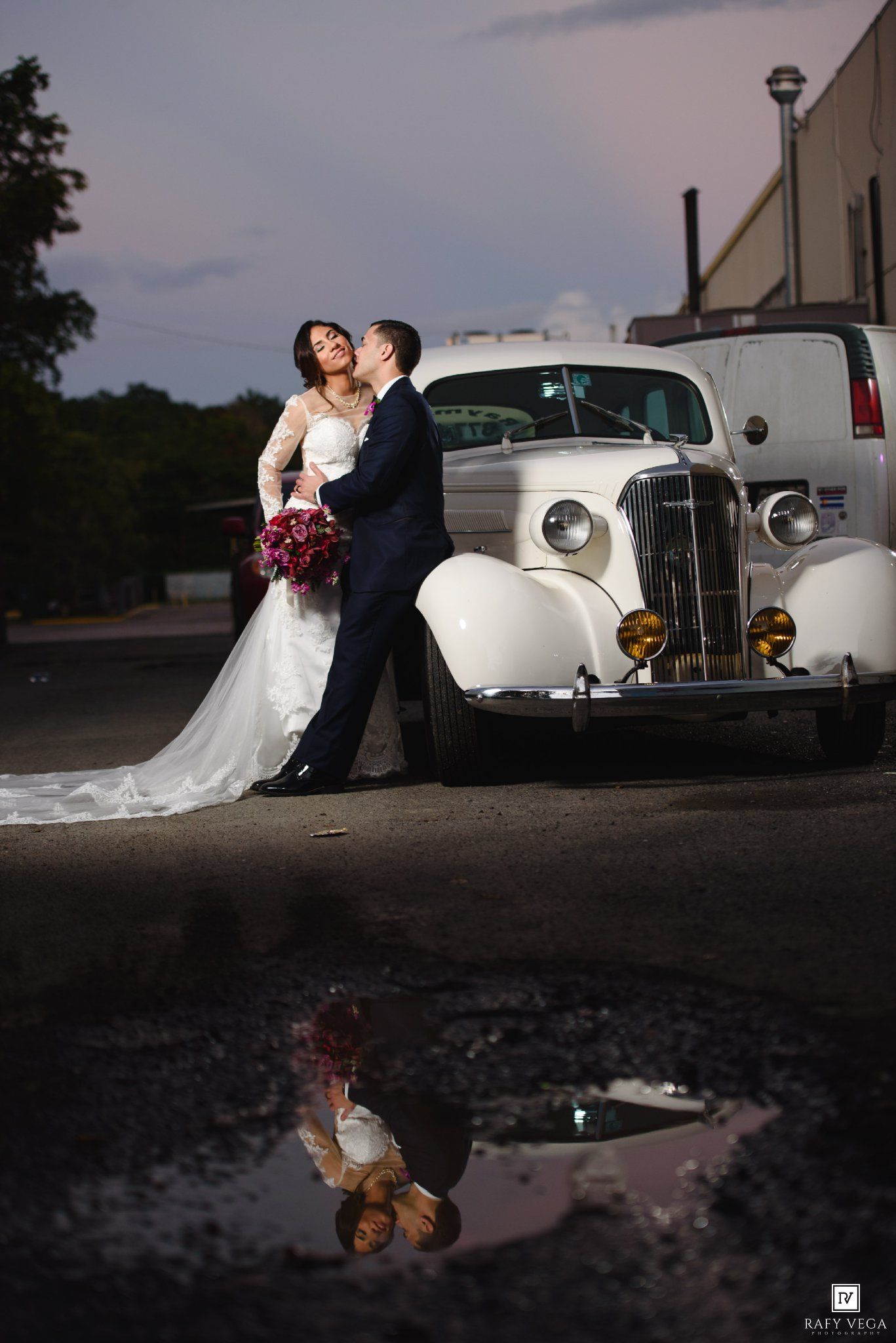 Bride and groom share a romantic kiss beside a classic white vintage car, with their reflection visible in a puddle below.