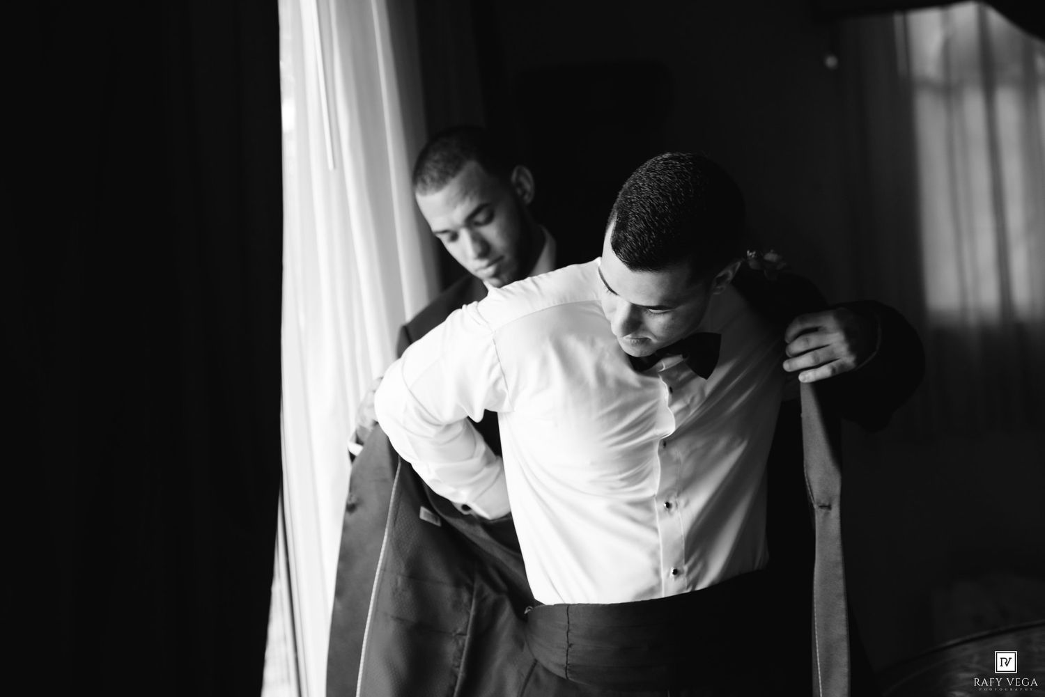 Black and white photo of a man helping another man put on a suit jacket near a bright window.
