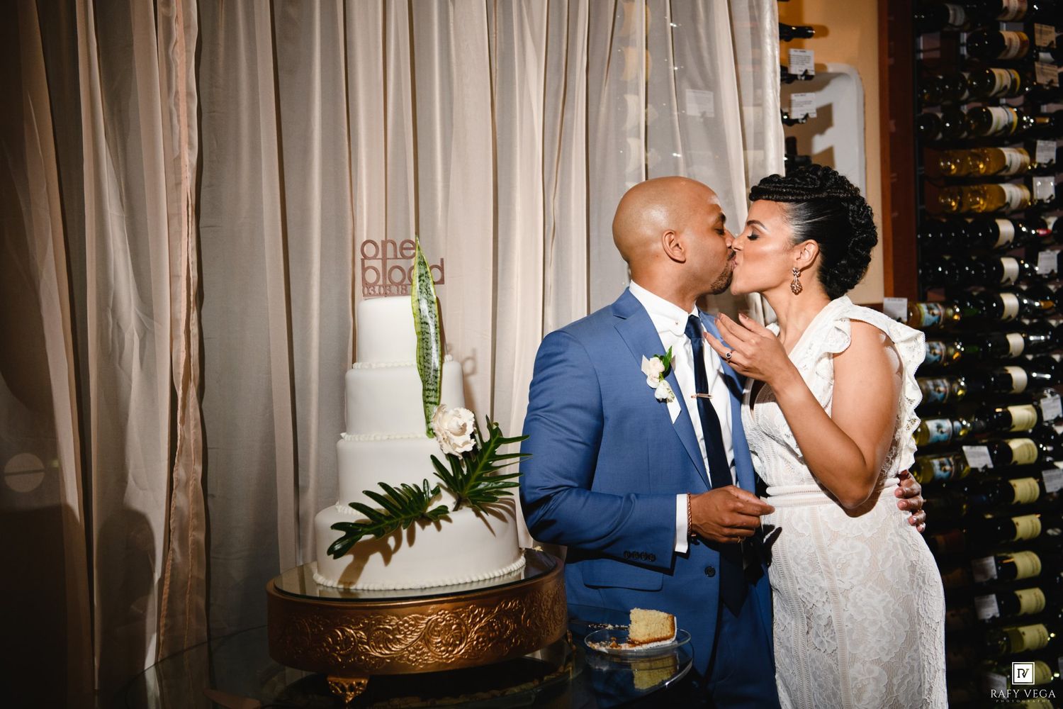 Newlyweds share a kiss beside a white wedding cake near a wine rack at their elegant reception venue.