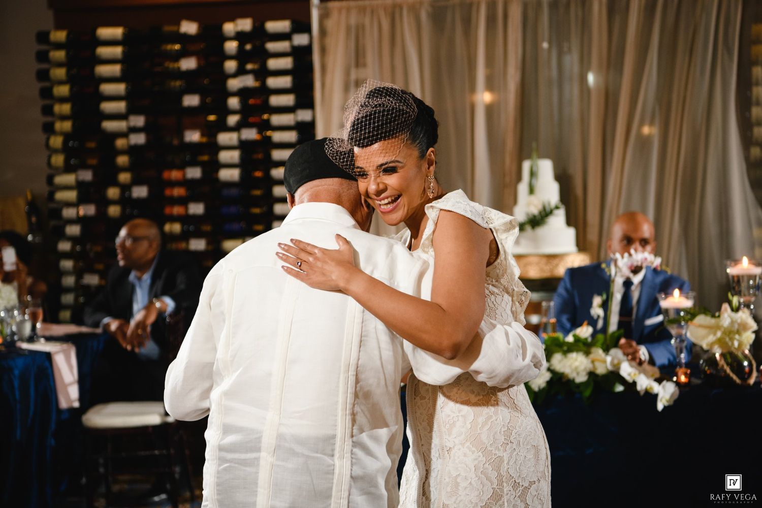 Bride in lace gown smiling and hugging a guest at elegant wedding reception with wine wall backdrop.