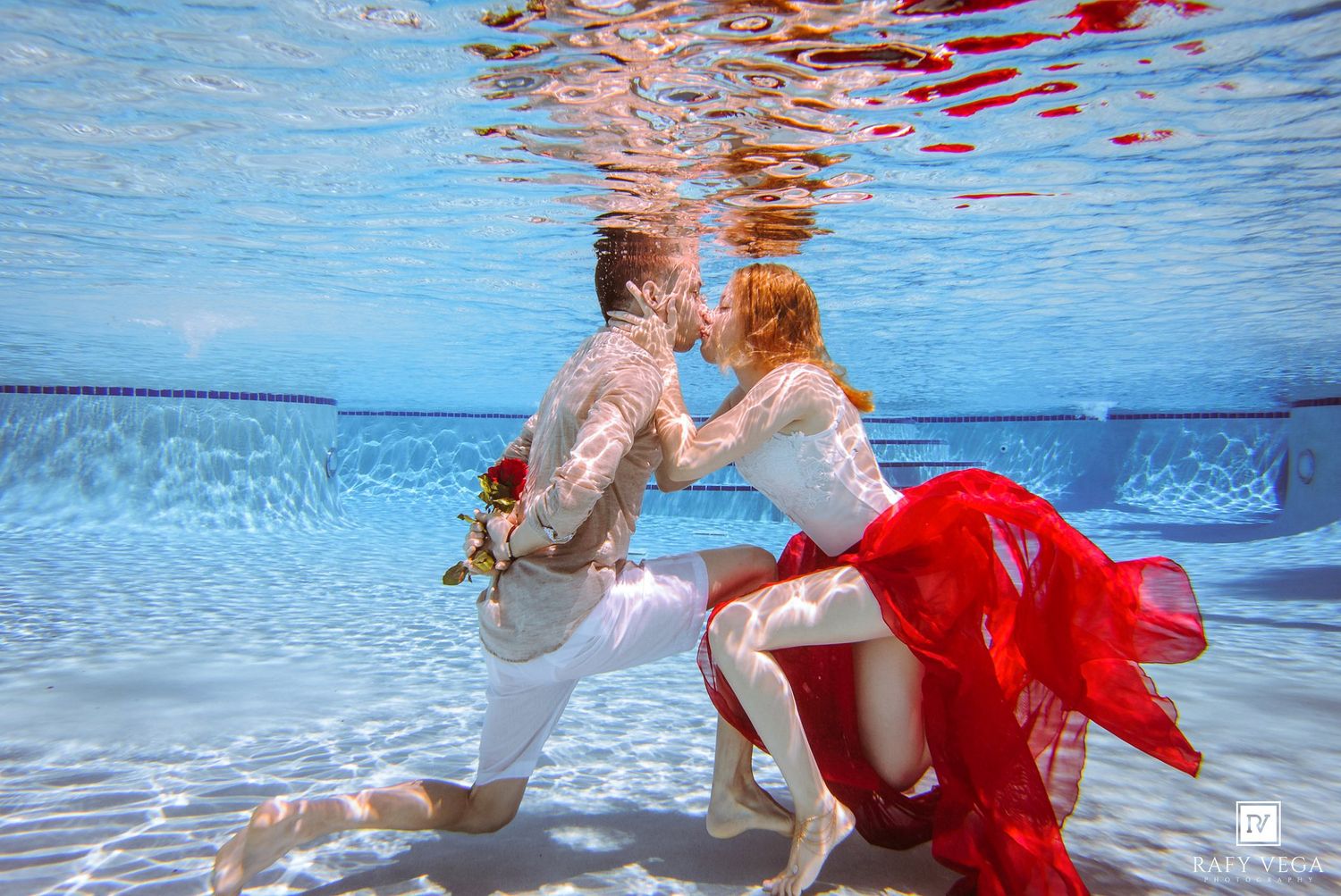 Couple sharing a romantic underwater kiss in a pool, woman in red flowing dress and white lace top, man in white outfit.