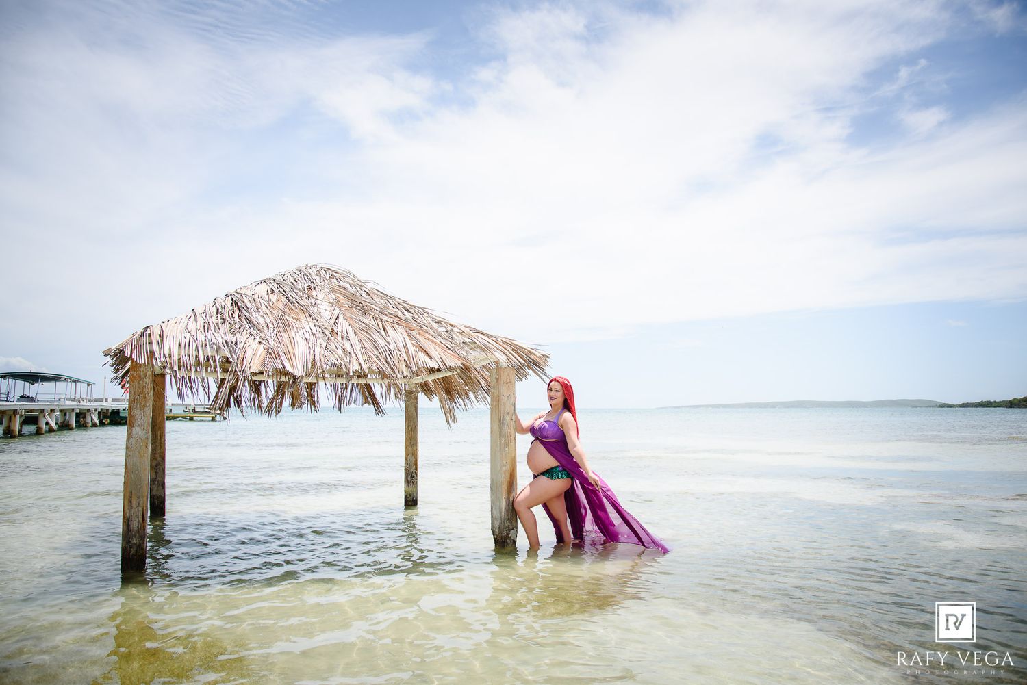 Pregnant woman in purple mermaid costume standing in shallow ocean water near a thatched hut.
