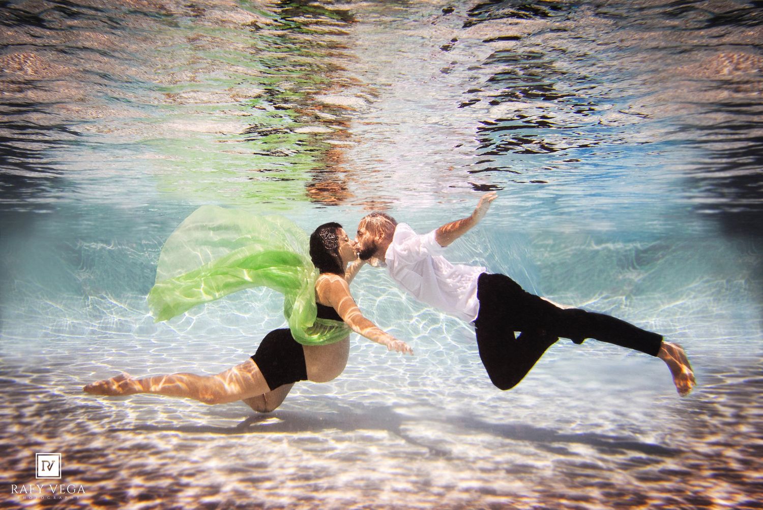 Couple poses underwater in a pool, woman in green flowing fabric and black outfit, man in white shirt and black pants.