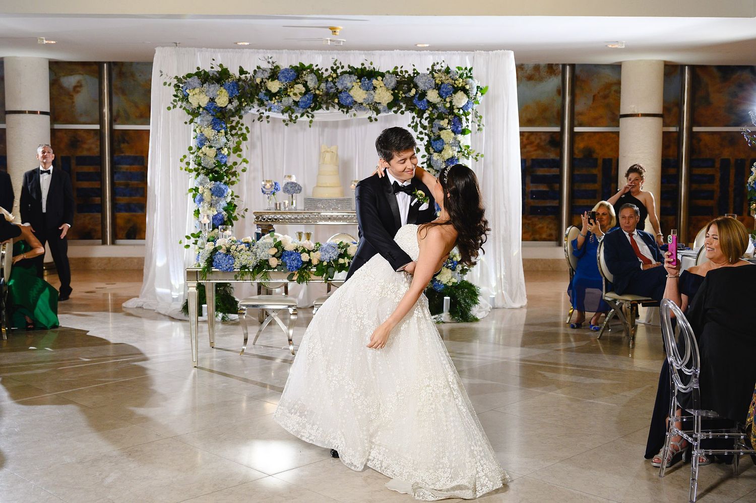 Groom dips bride during first dance at elegant wedding reception with floral arch backdrop.
