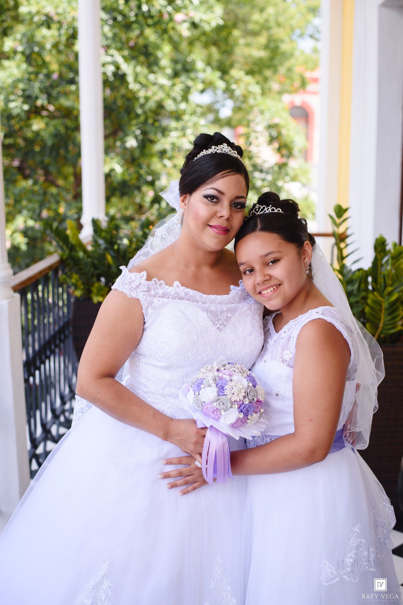 A woman and young girl in matching white wedding gowns with veils and tiaras pose together holding a purple bouquet.