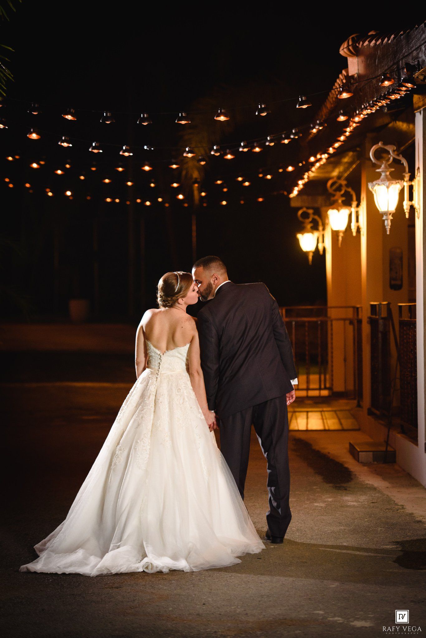 Newlyweds walk hand in hand under string lights outside a warmly lit venue on their wedding night.