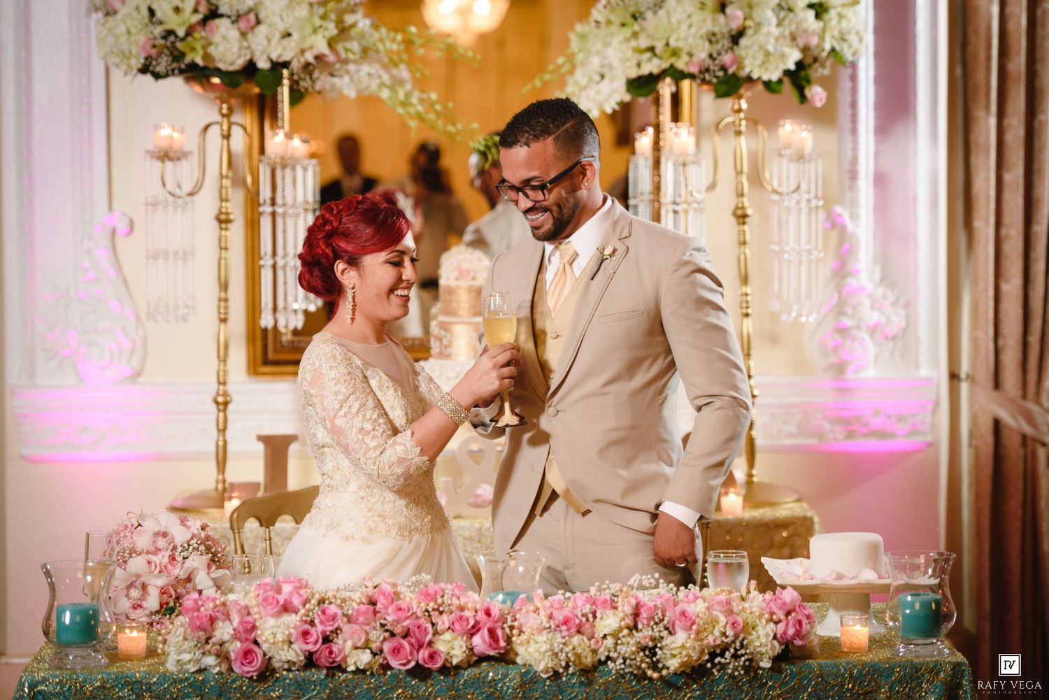 Couple sharing a toast at elegant wedding reception with pink floral arrangements and ornate gold candelabras.